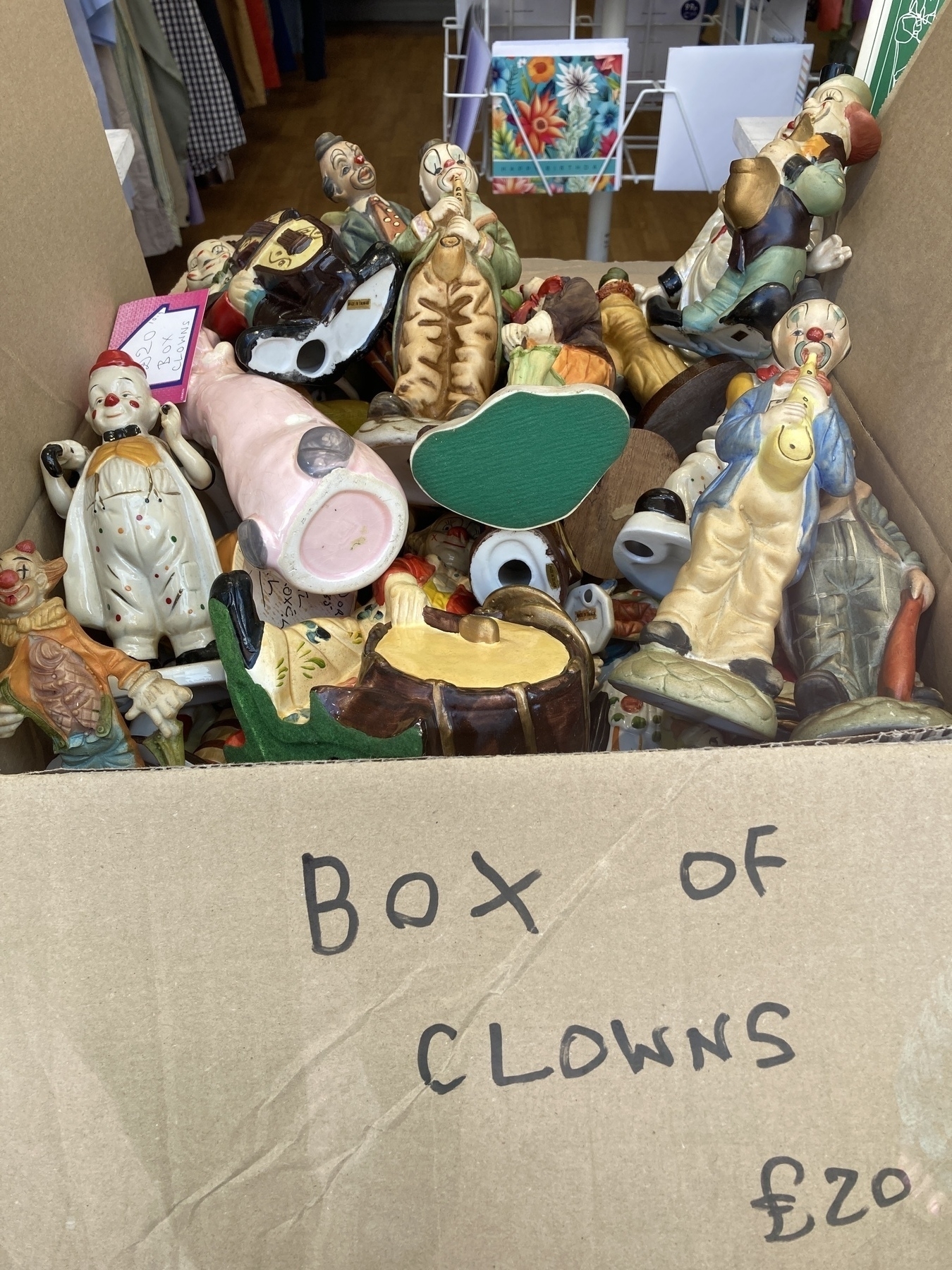 A box of assorted ceramic clown statuettes randomly piled. On the side of the is written “Box of clowns £20” in black marker. 